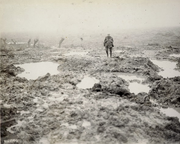 A black and white photograph of a lone soldier trudging towards the camera through the mud and puddles of a battlefield.