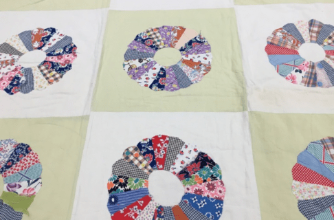 A portion of a bed quilt made of squares alternating in white and cream. Each square has a round "flower" shape made of patchwork pieces of colourful fabric.