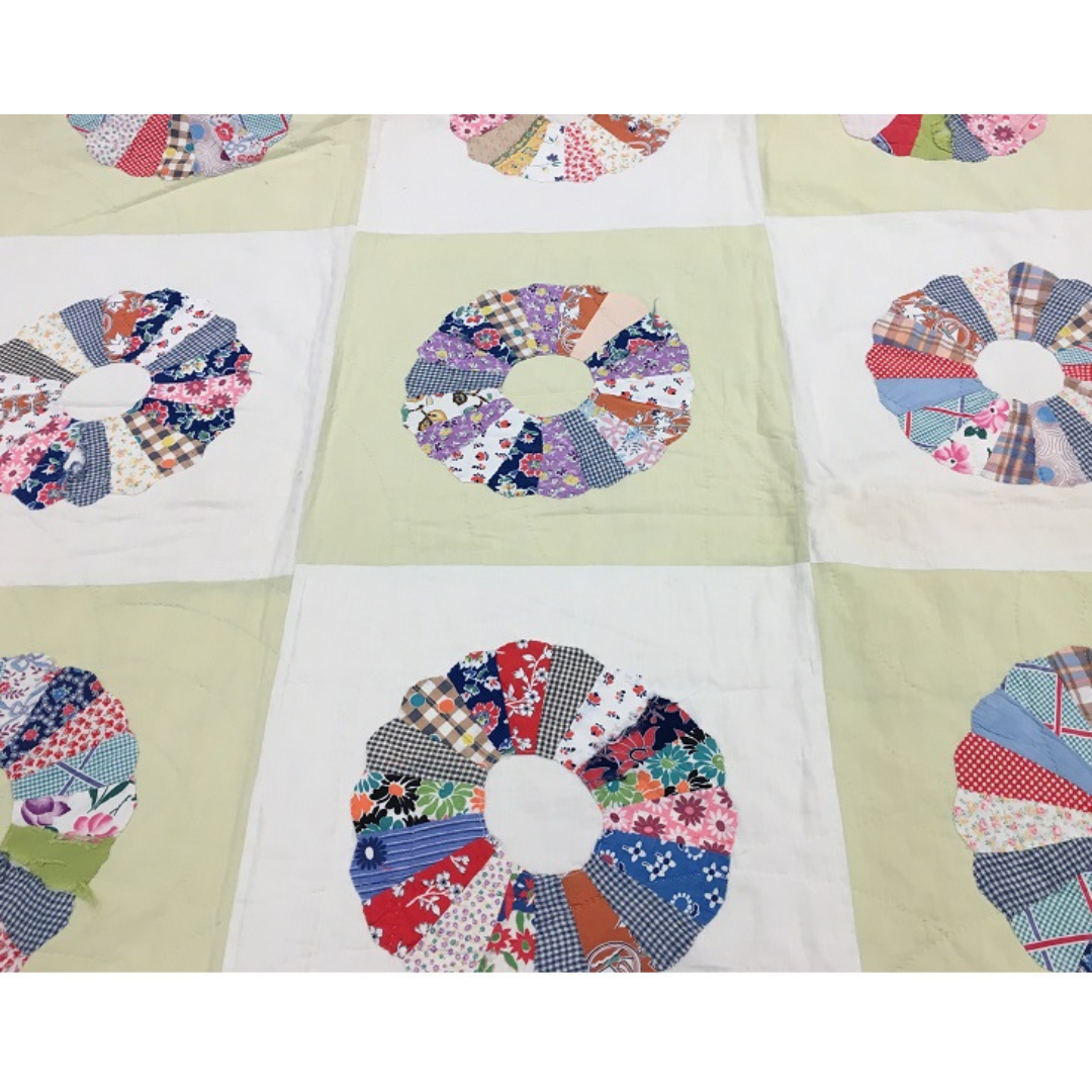 A portion of a bed quilt made of squares alternating in white and cream. Each square has a round "flower" shape made of patchwork pieces of colourful fabric.