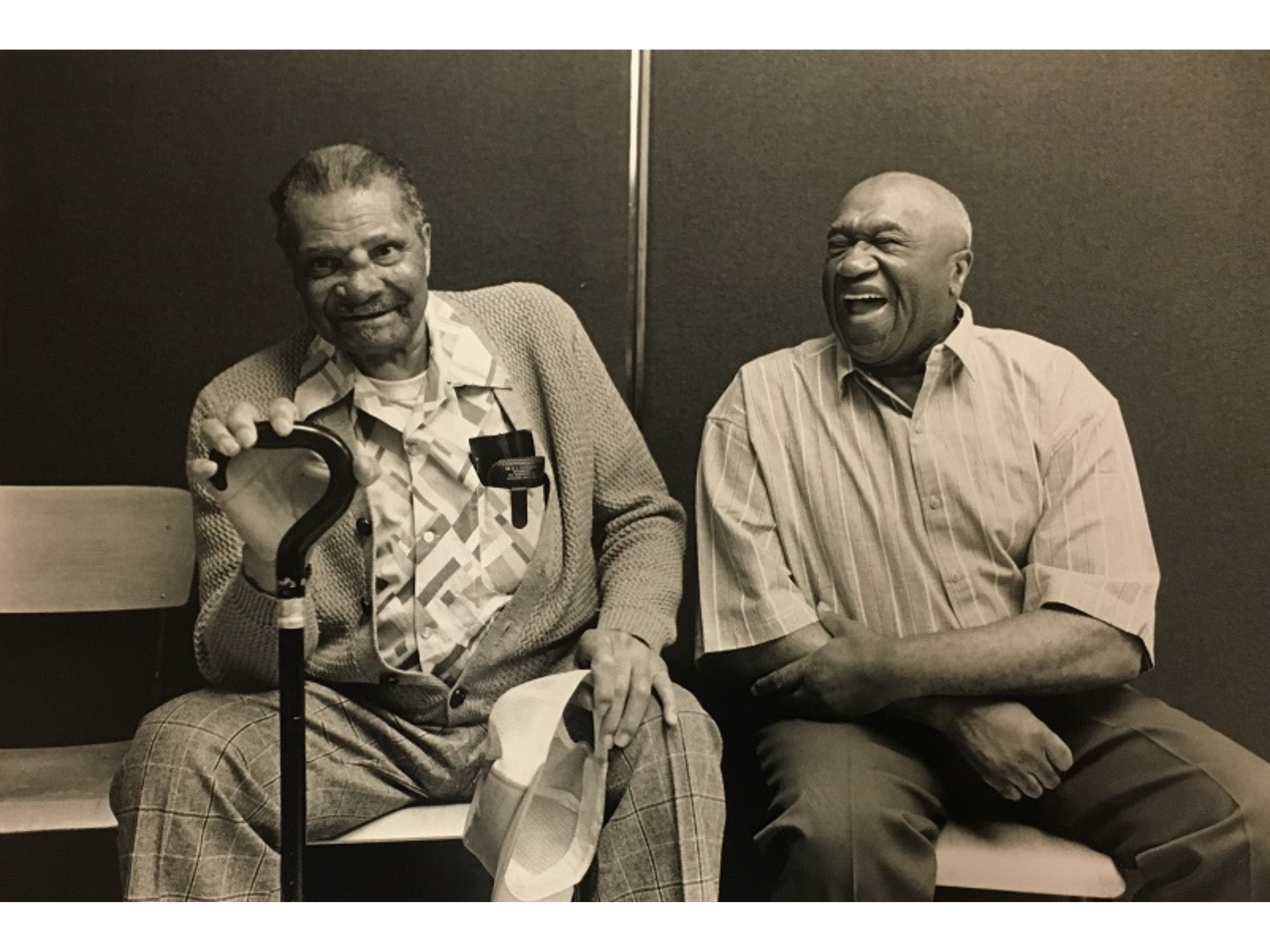 Two smiling Black men sitting together. One holds a walking cane.