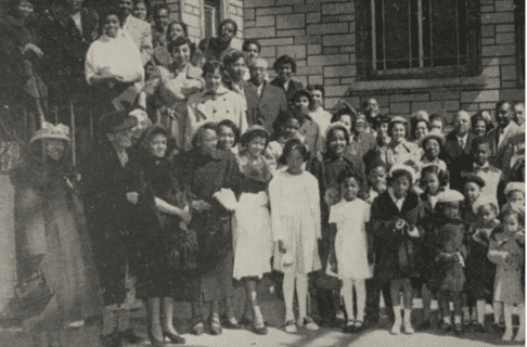 A black and white photograph of a large group of predominantly Black adults and children smiling outside of a brick building.