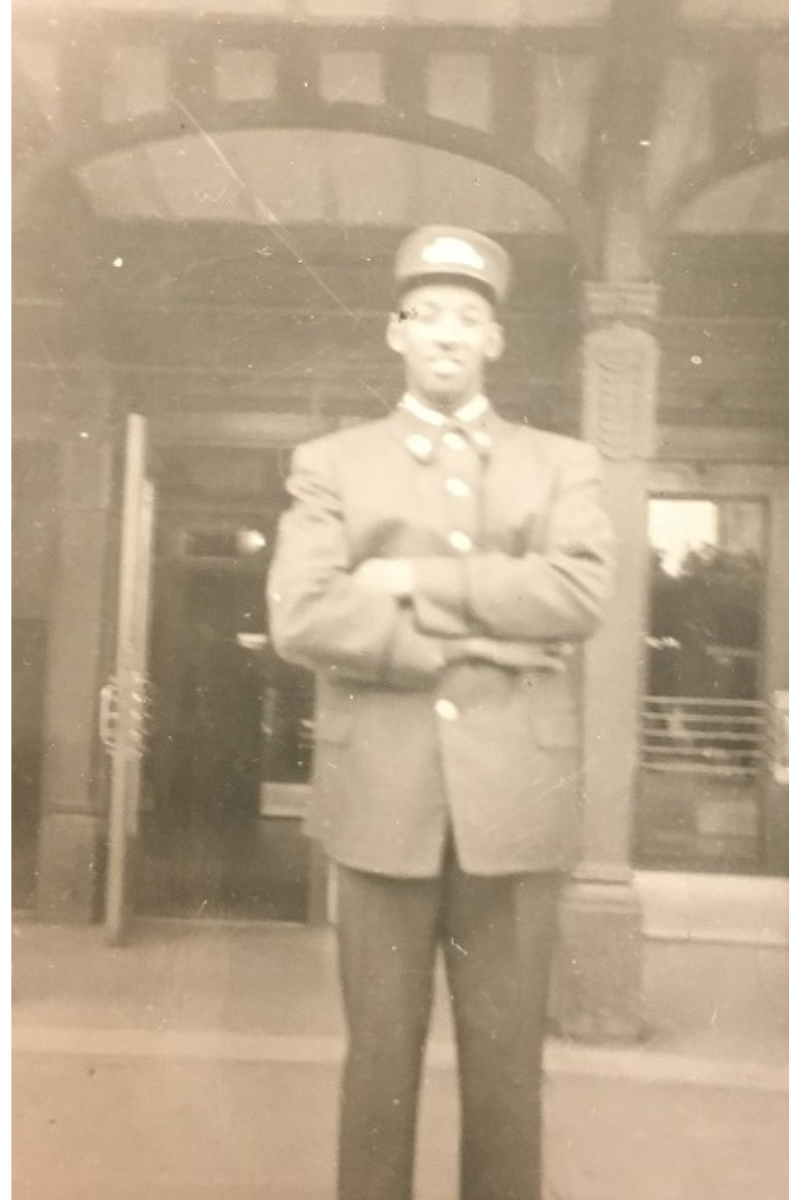 Sepia photograph of a smiling young Black man wearing a railway worker uniform. He is standing outdoors in front of a building entrance with his arms crossed in front of his chest.