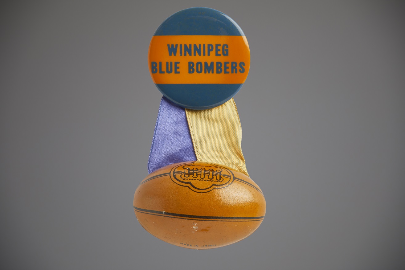 A Winnipeg Blue Bombers pin with a miniature football attached to a blue and orange button by blue and gold ribbons.