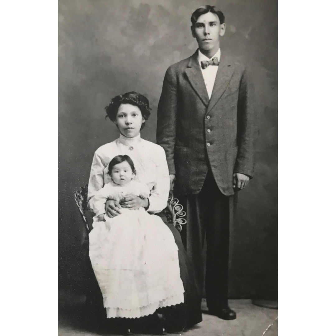 A black and white studio portrait of a young couple and baby in attire from the early 1900s. The young man stands beside the young woman who is seated with a baby on her lap.