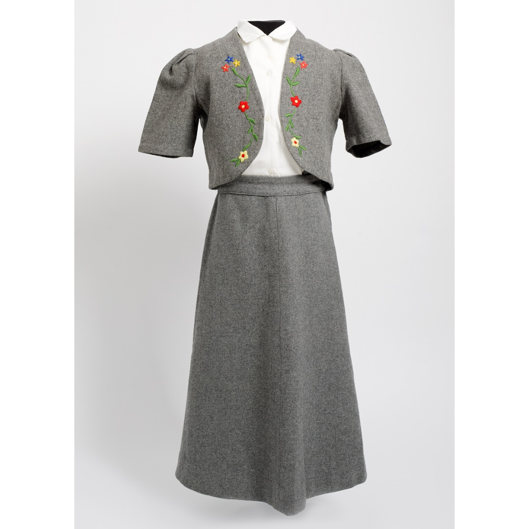 A child's size skit suit on a dress form. The skirt and jacket are made from a heathered grey material, with a few flowers embroidered on the lapels of the jacket. A button-up white shirt is underneath.