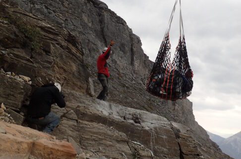 A large net suspended below a helicopter lifts crates of fossils and equipment off a precarious rock ledge.