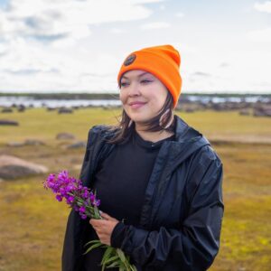 Photograph of a smiling individual standing in a grassy outstretch, looking off camera, wearing an orange toque and dark rain jacket, holding a small bouquet of purple-pink flowers.