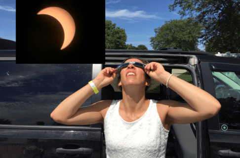 An individual standing outside of a car holds up a pair of eclipse glasses to their face as they look up at the sky. In the upper left corner is a photo of the sun mid-solar eclipse.
