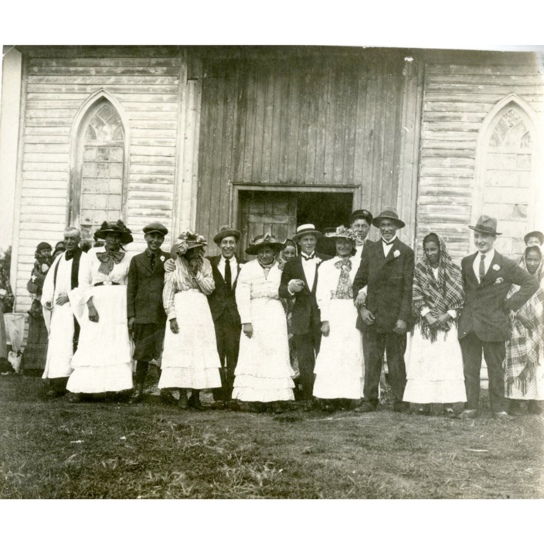 Sepia-toned photograph of a group of men and women standing outside of a wooden church building. The women are wearing light coloured dresses and hats, and the men are wearing suits and hats. A few in the group are smiling or laughing.
