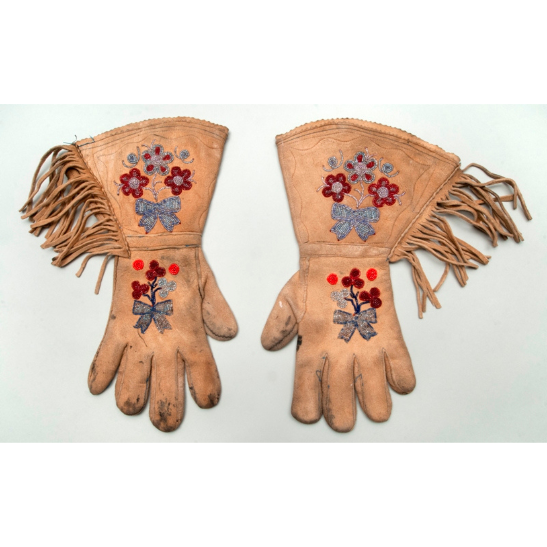 A pair of worn light tan gauntlet gloves with flower and berry beadwork on the backs of the hand and wrist portion, and a fringe along the outside of the wrist.