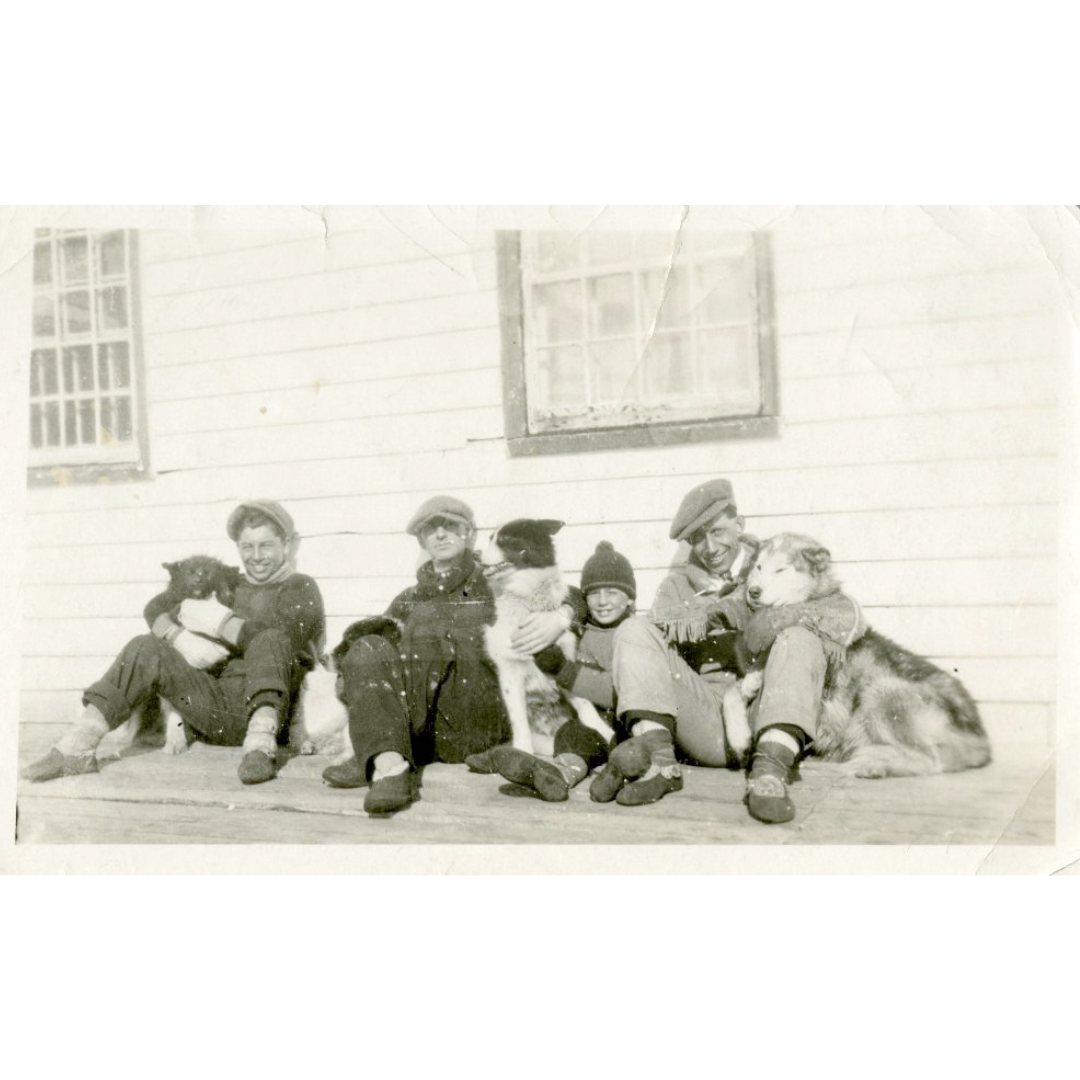 Three men and a child sit outdoors against the outer wall of a building, wearing hats and coats, along with three seated dogs.