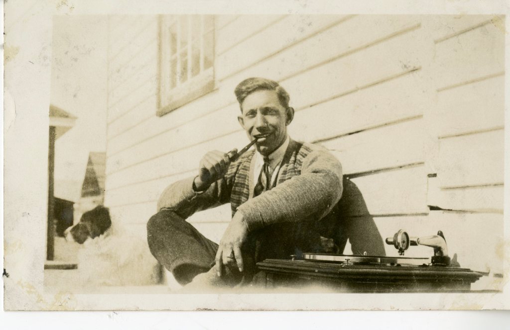 Sephia-toned photograph of a man seated outside, leaning against a building with his legs drawn up, with a pipe in his mouth.