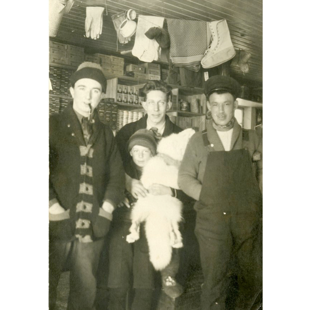 A sepia toned photograph of three men and a child smiling at the camera. They are standing indoors, with shelves stocked with supplies behind them, and a laundry line of mitts, boot liners, and other clothing items hanging from the ceiling above them.