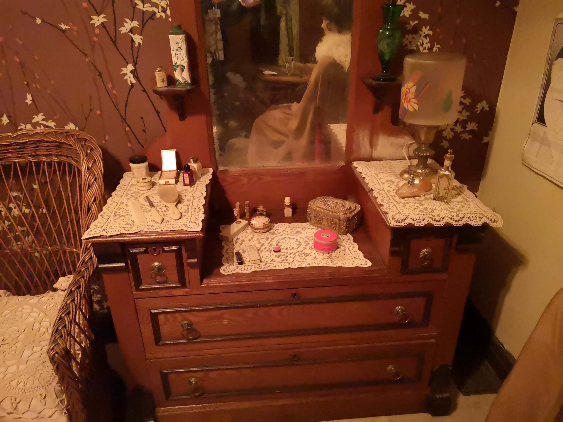 A wood dressing table with three levels. Each has a doily on it and various personal items including make up, a brush, perfume, and a jewelry box.