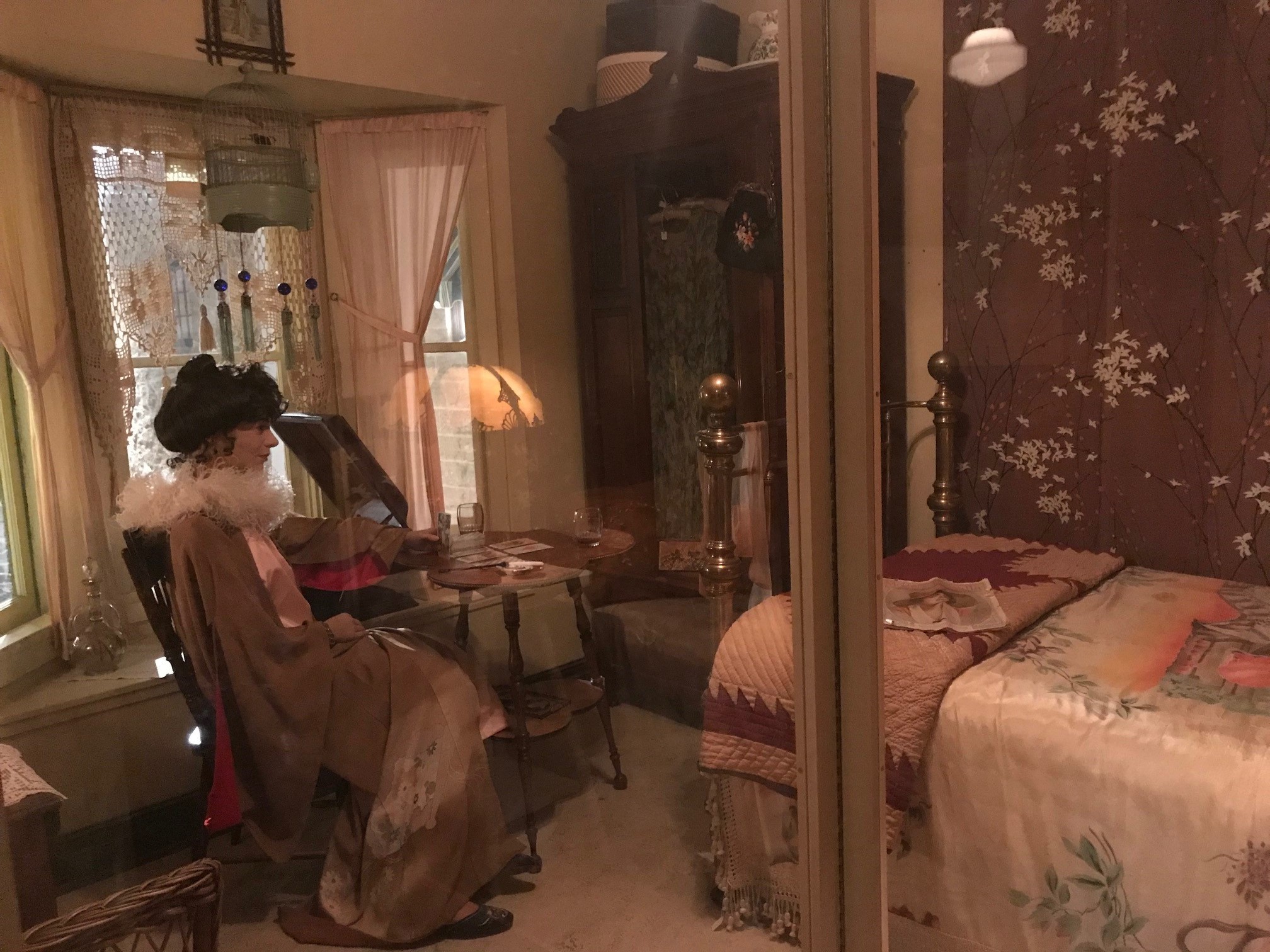 A bedroom. A mannequin of a woman from behind wearing a robe and ruff is seated at a small table with tarot cards laid out on it near a bay window. On the right side of the room is a small neatly-made bed with a quilt folded along the foot. A wardrobe is in the corner across from the centre of frame.
