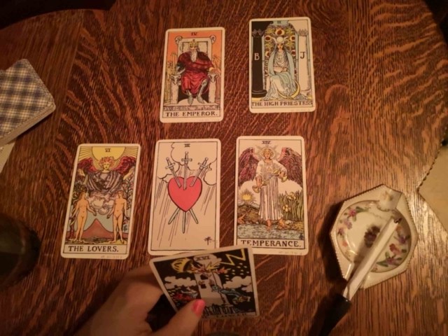 Looking down onto the surface of a table where four tarot cards are laid out surrounding a face-down deck: the Emperor, the High Priestess, the Lovers, and Temperance. At the bottom a hand holds a yet-to-be-placed card: the Tower. To the right of the cards is a small ashtray and cigarette holder.
