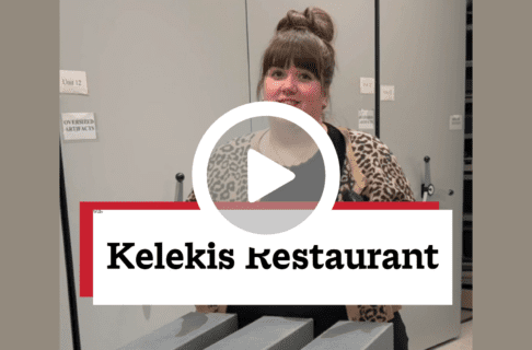 A screenshot of a video, an individual standing in a storage room in a hall between rows of cabinets, smiling towards the camera. There's a play button over the screenshot and overlaid text reads, "Kelekis Restaurant".