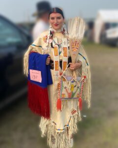 A young woman wearing a traditional Anishinaabe dress holding up a beaded blanket, bandolier bag, and fan.
