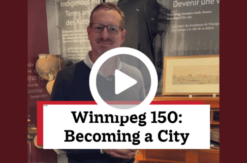 A screenshot of a video, an individual standing in front of a display case containing a number of artifacts including a large desk. There's a play button over the screenshot and overlaid text reads, "Winnipeg150: Becoming a City".
