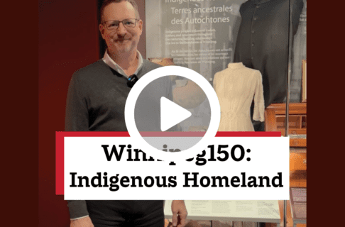 A screenshot of a video, an individual standing in front of a display case containing a number of artifacts including a jacket and a white dress. There's a play button over the screenshot and overlaid text reads, "Winnipeg150: Indigenous Homeland".