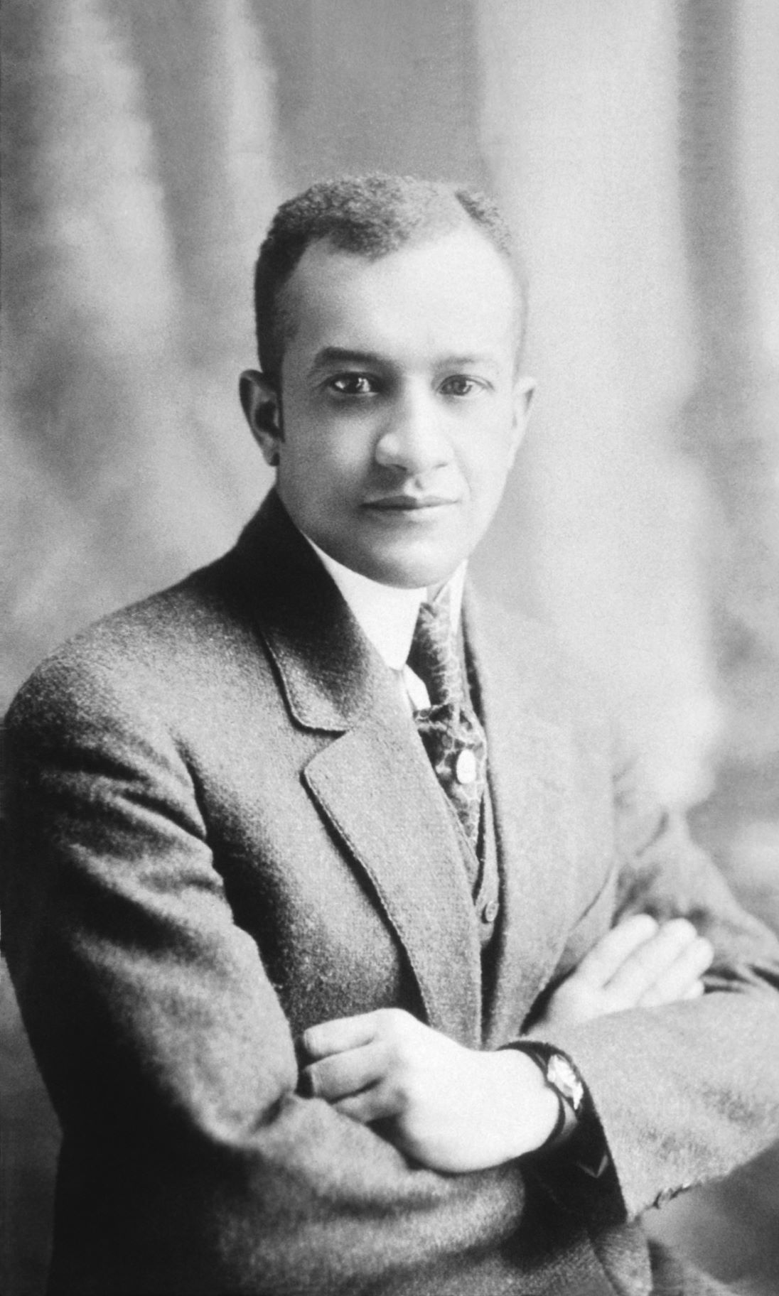 A black and white photograph of a young Black man posed with his arms crossed in a studio portrait. He is wearing a suit and tie.