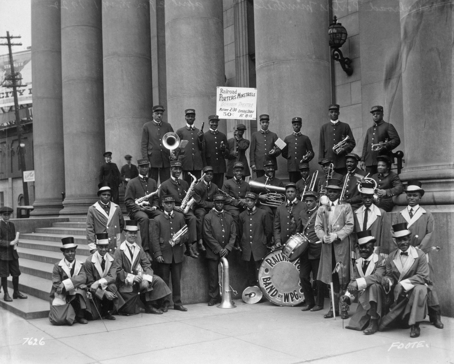 A black and white photograph of a group of Black men in uniform posing for the photo. Many are holding instruments like trumpets, saxophones, tubas, or drums, and some carry batons. In the centre back, one holds up a sign that reads, "Railroad Porters Minstrels / Dominion Theatre".