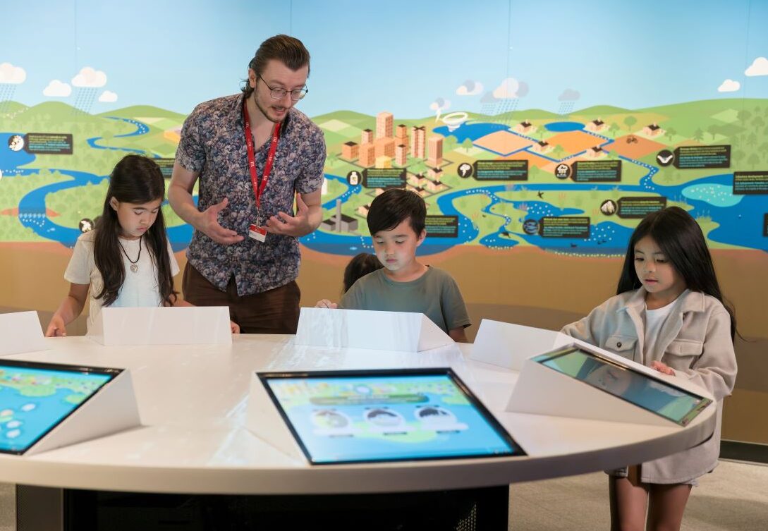 An adult and three children engage with digital exhibit screens on a round table. A mural showing the water system is on the wall behind them.