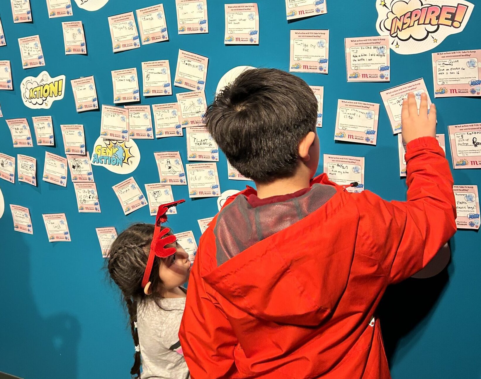 Two children placing sticky notes on a blue wall filled with other previously placed notes.
