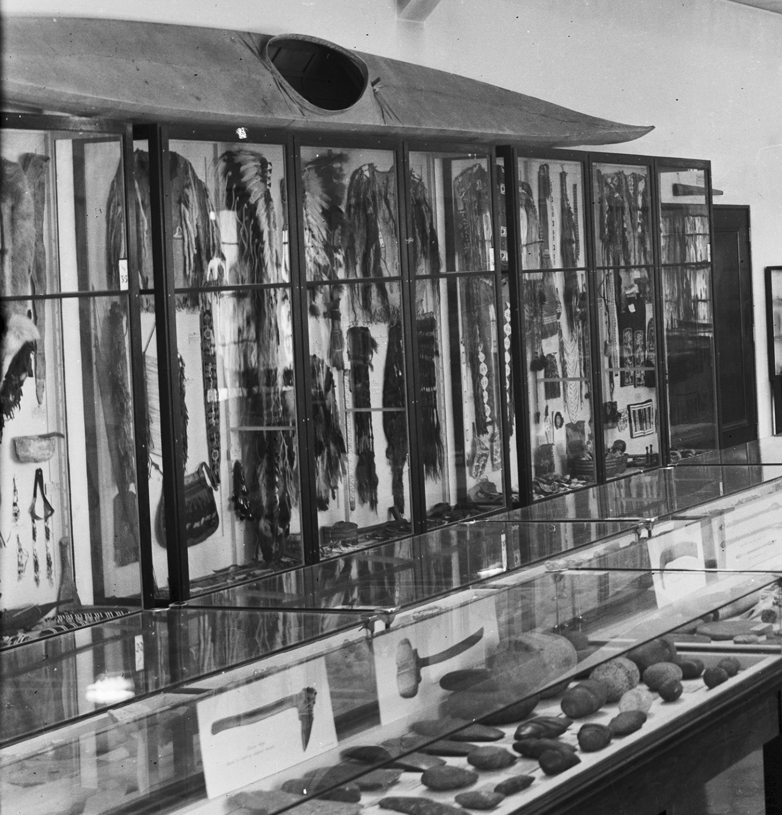 Black and white image of museum display cases showcasing various Indigenous artifacts, including a kayak. 