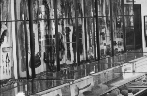 Black and white image of museum display cases showcasing various Indigenous artifacts, including a kayak.
