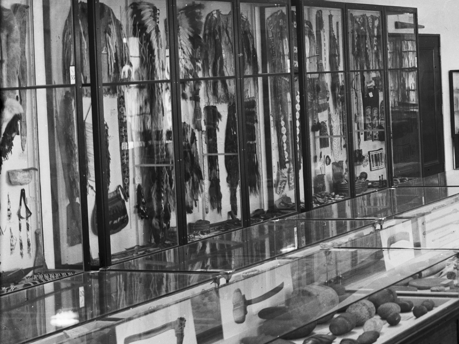 Black and white image of museum display cases showcasing various Indigenous artifacts, including a kayak.