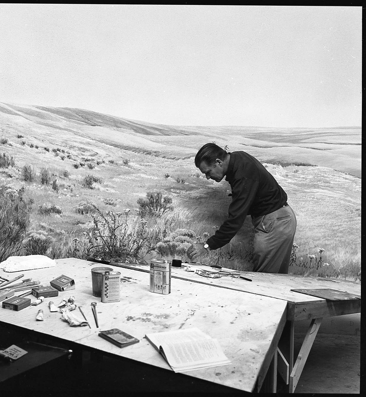 Black and white image of a man painting a mural of a gently-hilled prairie landscape; table with various art supplies in the foreground.