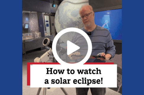 A screenshot of a video, an individual standing in front of a model of the moon next to a telescope, holding a pair of eclipse glasses. There's a play button over the screenshot and overlaid text reads, "How to watch a solar eclipse!".
