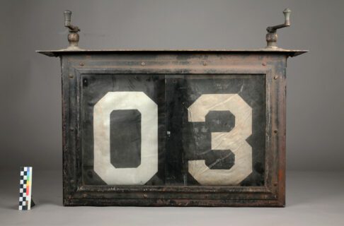 A somewhat worn looking rectangular sign with cranks on either end of the top. On the face are two fabric strips showing the numbers 0 and 3.
