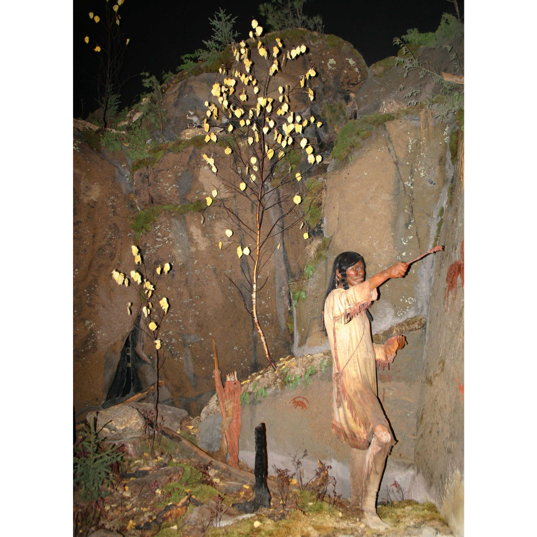 Looking into a Museum diorama where a mannequin is posed painting a cliff wall next to autumn trees.