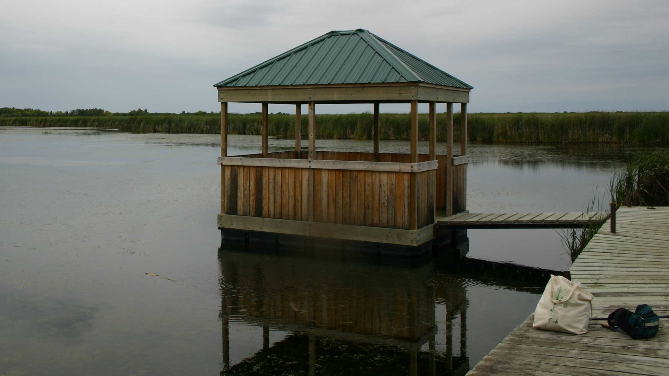 A floating wooden view point at the end of a boardwalk on a lake on a cloudy day.