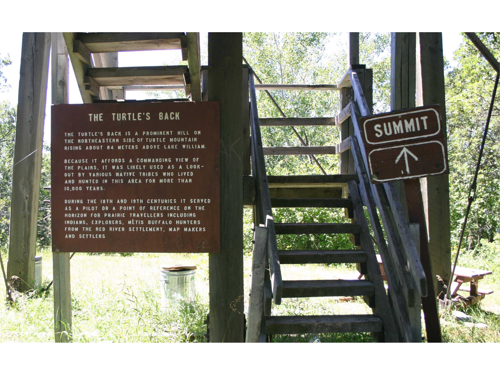 Standing at the base of a set of wooden outdoor stairs. Either side of the staircase are wooden signs. One reads "Summit" with an arrow pointing up, and the other is an information panel about "The Turtle's Back".
