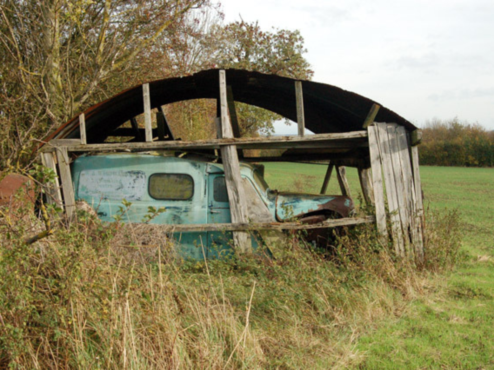 A derelict light-blue vehicle in a derelict garage that is more open than covered at this point.