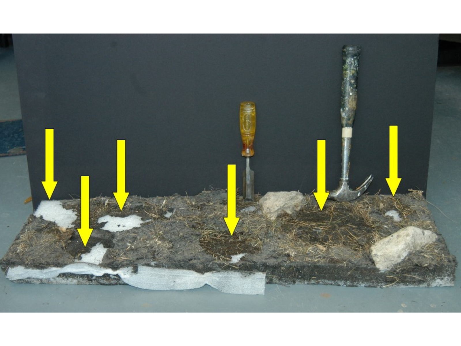 The same portion of diorama base as the previous two image, now looking even barer. In places the white ethafoam base is visible through the surrounding ground and grass areas, signified with yellow arrows. Propped up at the back of the piece are a flathead screwdriver and a hammer.