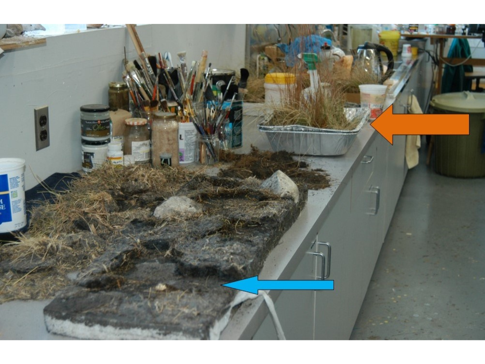 The same diorama portion as the previous three images on a countertop from the side. The white portions of foam have been painted to match the dirt (signified by a blue arrow), and in an aluminum tray sits several clumps of grasses (signified with an orange arrow).