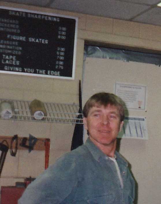 A grainy colour-film photograph of a young blonde man wearing a blue denim shirt standing in the workshop area of Vimy Arena.