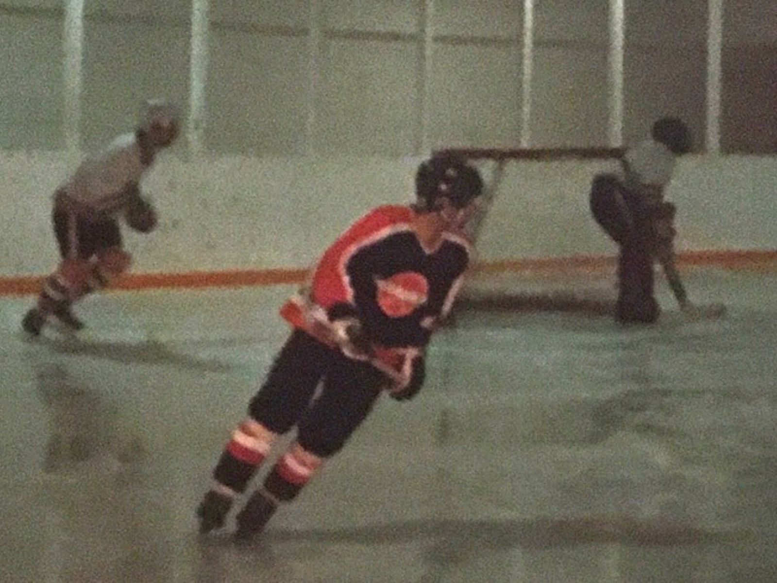 A pixelated colour-film photograph showing three hockey players on the ice of an indoor rink. The player in the centre, Allan Merko, is wearing a dark coloured jersey with red accents and a black helmet.