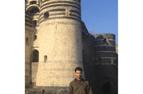 A young man in a brown windbreaker standing in front of a stone tower.