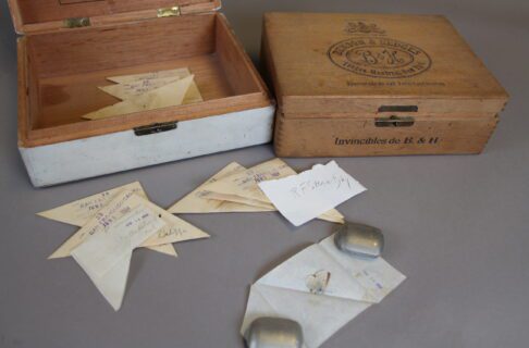 Two wooden boxes, one of which has the hinged lid open, with triangle folded papers inside and in front of the box. One of the papers is unfolded, and held open by paperweights, revealing a preserved butterfly specimen inside.