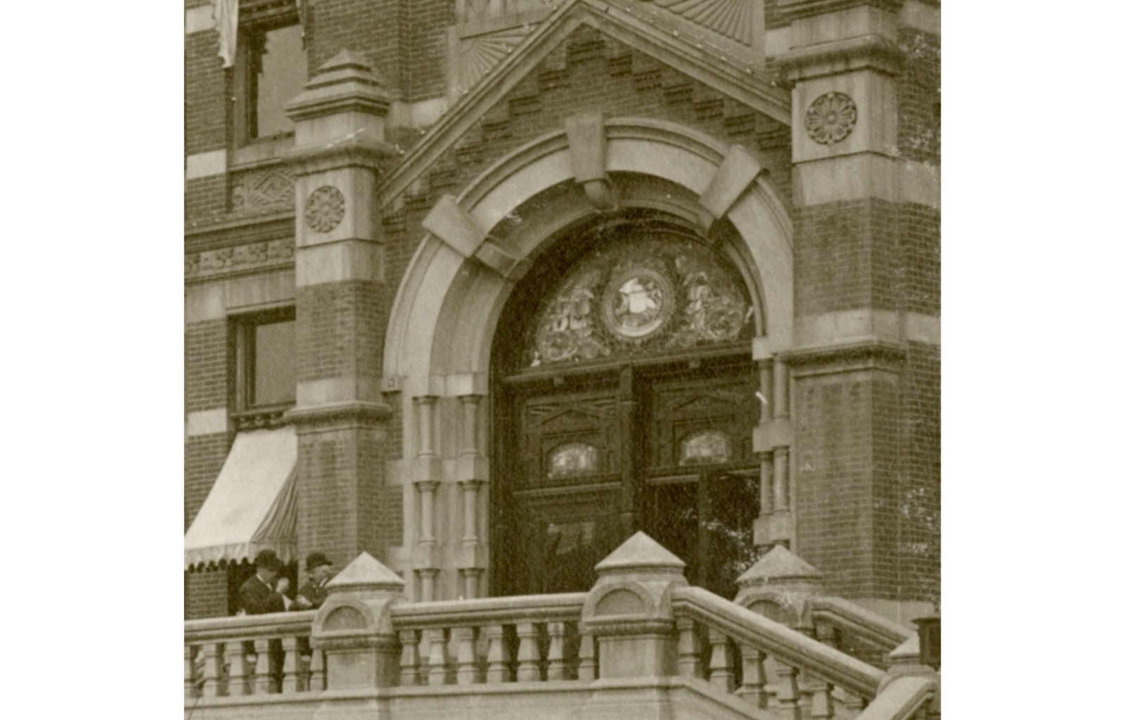 Sepia-toned photograph of the entry way to a brick building. Over elaborately carved doors sits a semi circle stained glass window below a doorway-framing arch.