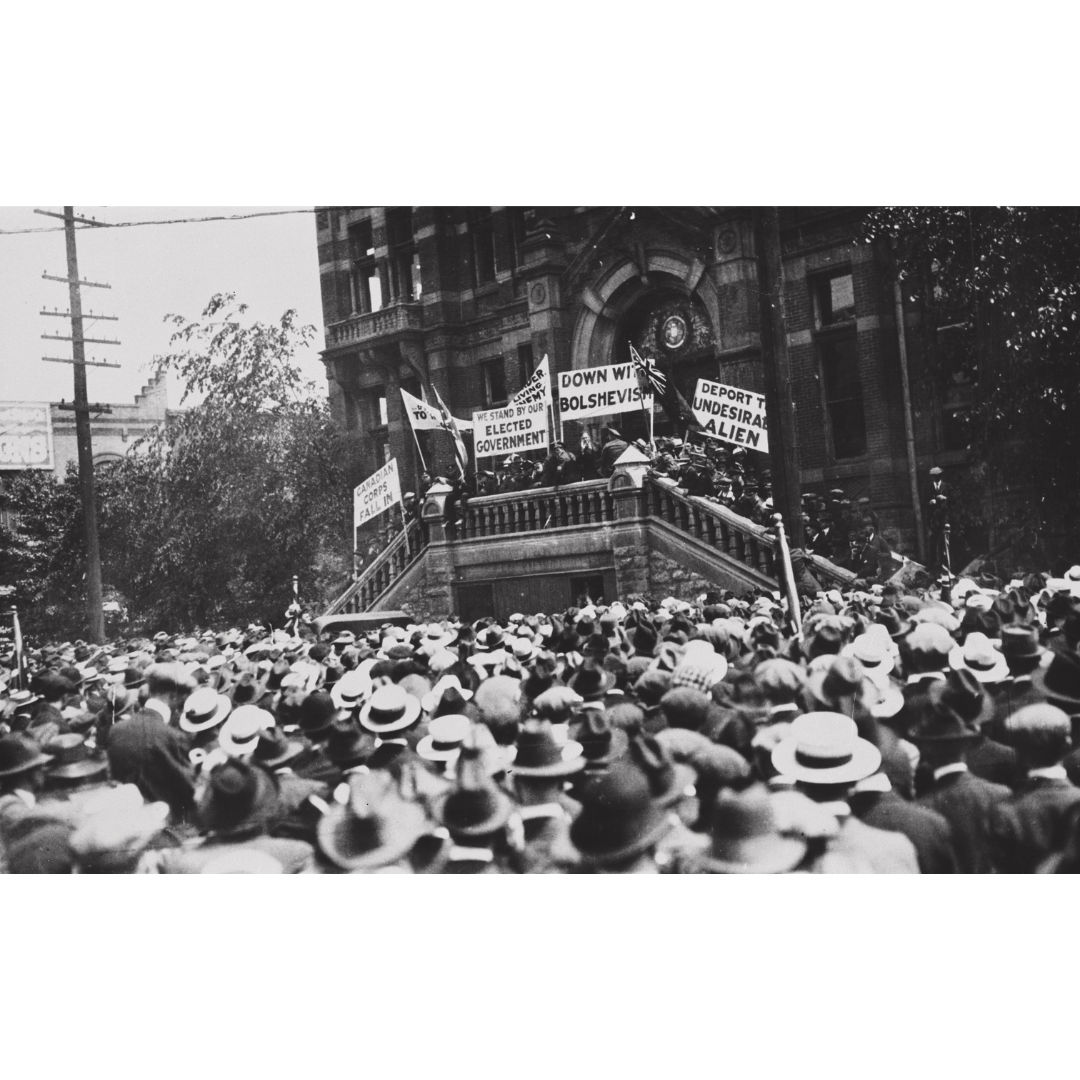 A black and white photograph looking our over a crowd gathered in front of a Victorian-style brick building. Groups standing on the entrance staircase and balcony hold up signs reading things like, "Canadian Corps Fall in", "We stand by our elected government", "Down with Bolshevism", and "Deport the undesirable alien". Above the door behind them is a semi-circle stained glass window.