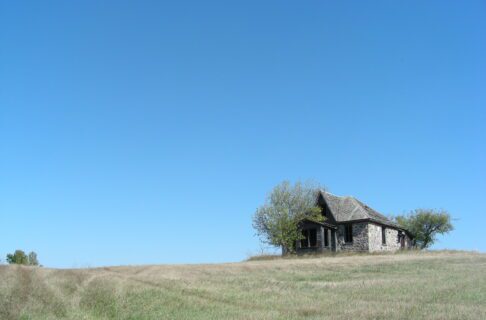 An empty old stone house on the top of a grassy knoll, against a blue sky. Trees grow wild close to the walls of the house.