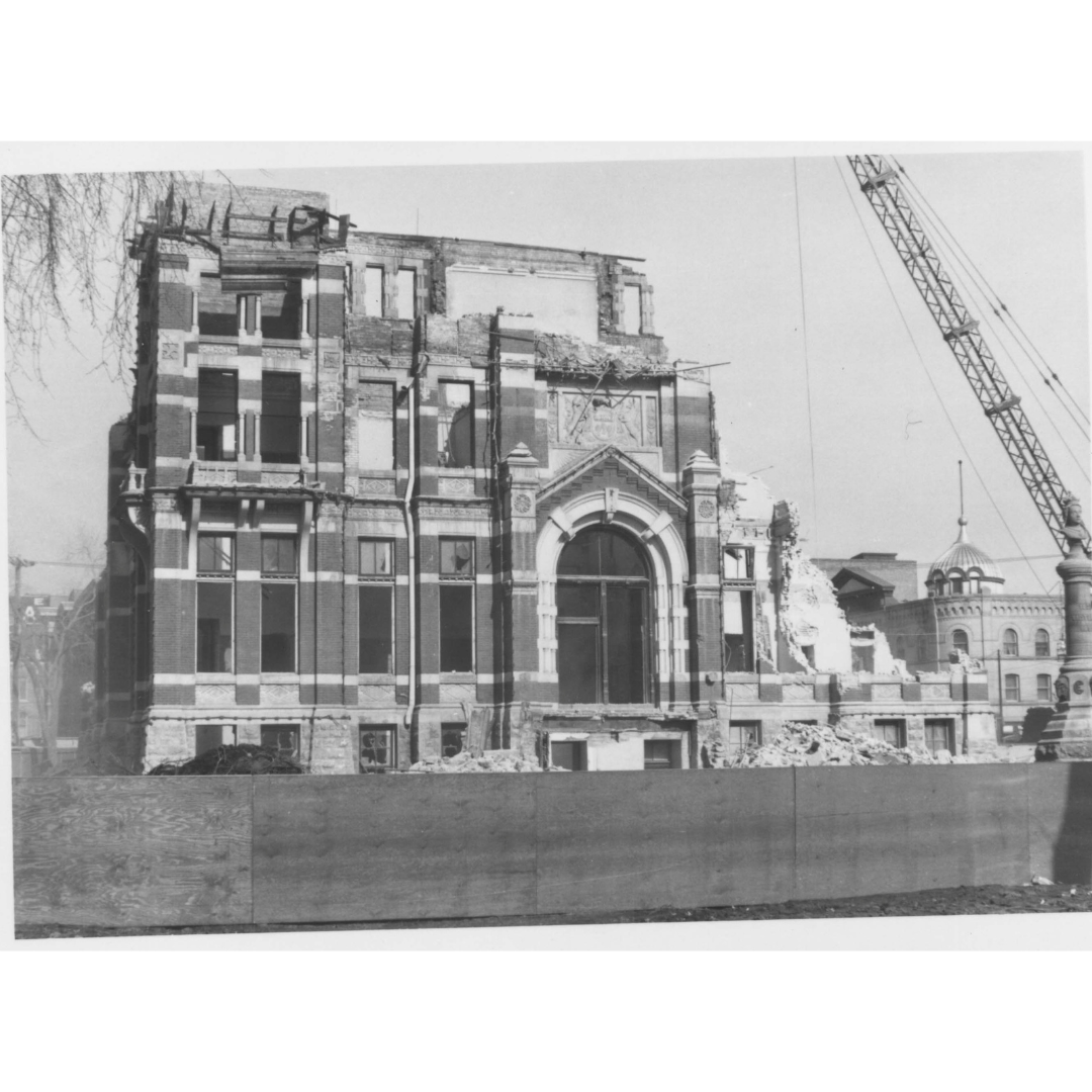 A black and white photograph of a partially demolished brick building. On the left side, the lower portions of the walls still stand, but the right side is further removed.
