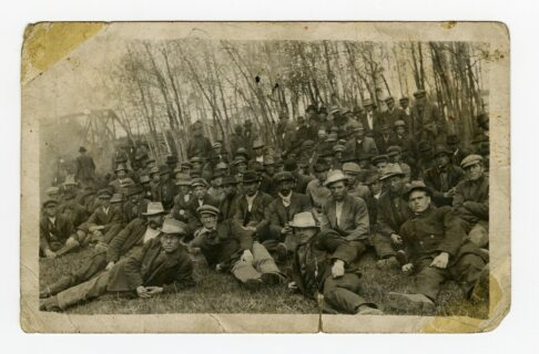A worn sepia-toned photograph showing a group of men posing for a photograph. Many sit or lounge on the grass, and a few stand at the back. they are all wearing 1910s work wear and hats.