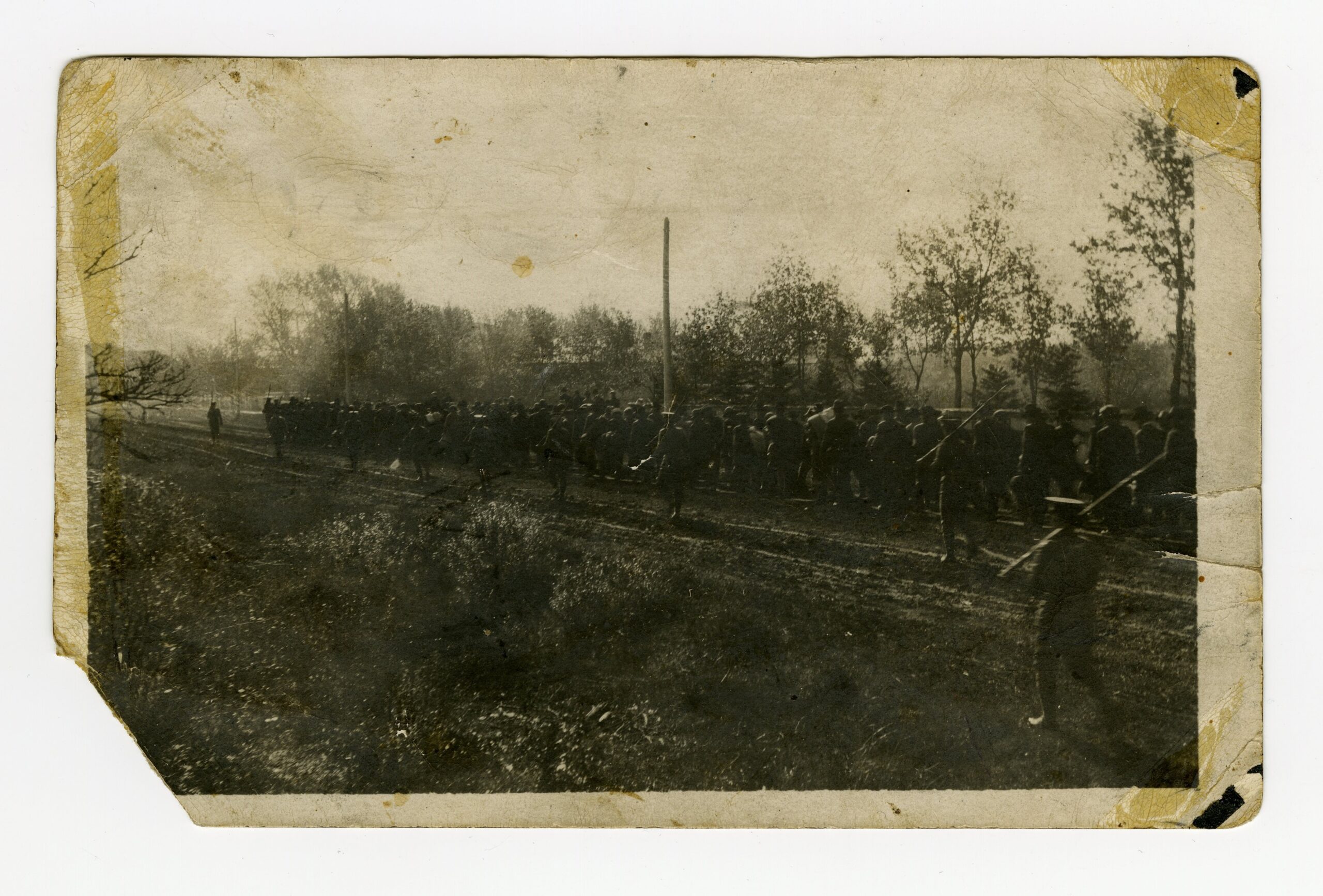 A worn sepia-toned photograph with ths bottom left corner torn off, showing a group of men marching into the distance, escorted by individuals with rifles at attention on their shoulders.